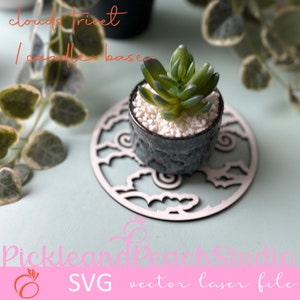 Puede incluir: Un salvamanteles de madera blanco con un diseño de nubes, con una pequeña planta suculenta en una maceta azul. El salvamanteles está etiquetado como "clouds trivet candle base".
