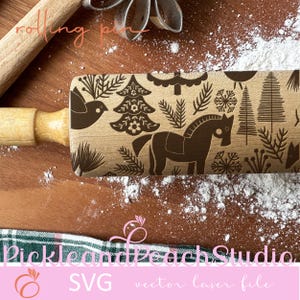 Puede incluir: Un rodillo de madera con un diseño marrón y blanco de un caballo, árboles, pájaros y copos de nieve. El rodillo está sobre una superficie de madera con polvo blanco sobre él. El texto "Pickle and Peach Studio SVG vector laser file" está sobre un fondo rosa debajo del rodillo.