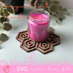 May include: A pink candle in a glass jar sits on a honeycomb-shaped wooden trivet. The trivet is made of cork and has a natural wood finish. The text "Honeycomb Trivet Candle Base" is written above the candle.