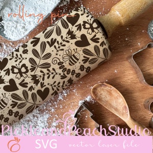 Könnte beinhalten: Ein Holz-Nudelholz mit einem braunen Blumenmuster, Herzen, Schmetterlingen und Bienen. Das Nudelholz liegt auf einer Holzoberfläche mit Puderzucker und Ausstechformen.