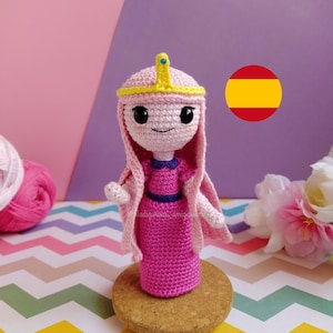 Op de afbeelding: Handgemaakte gehaakte pop van Princess Bubblegum uit Adventure Time. De pop is voornamelijk roze, met een gele kroon en een paarse accent op de jurk. Hij staat op een kurken voet.