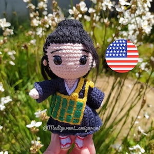 Puede incluir: Muñeca amigurumi de crochet con un atuendo tradicional coreano azul y verde. La muñeca tiene cabello negro y grandes ojos marrones. La muñeca sostiene una pequeña bandera estadounidense.