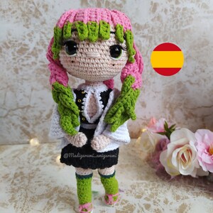 May include: A crocheted doll with pink and green hair, wearing a white and black dress. The doll is standing on a white surface with a blurred background.