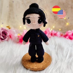 May include: A handmade crochet doll with black hair styled in a bun and wearing a navy blue outfit. The doll has large, black eyes and is standing on a cork base. The doll is a unique, handcrafted item.