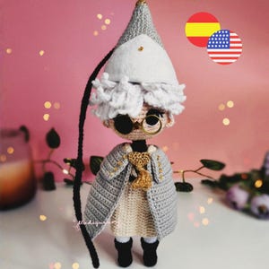Puede incluir: Una muñeca de ganchillo hecha a mano con pelo blanco rizado, un sombrero gris alto y puntiagudo y gafas redondas con montura dorada. La muñeca lleva un vestido beige, un abrigo gris con detalles dorados y botas negras.