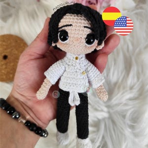 Puede incluir: Muñeca de ganchillo hecha a mano, cabello negro, camisa blanca con botones dorados, pantalones negros y zapatos blancos. El texto "MADIGURUMI" es visible. Una pulsera está en el fondo.