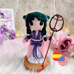 Puede incluir: Muñeca amigurumi hecha a mano con cabello verde, vestida con un atuendo rosa y morado, y sosteniendo una pequeña red. La muñeca está tejida a ganchillo y se encuentra sobre una base de corcho. Los ojos de la muñeca son grandes y negros.