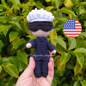 May include: A crocheted amigurumi doll wearing a blue suit, a white hat, and a black eye mask. The doll is being held in a hand with the American flag in the background. The doll's Instagram handle is @Madigurumi_amigurumi.