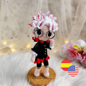 May include: A handmade amigurumi doll with white hair streaked with red, wearing round glasses and a black outfit with red accents. The doll is standing on a cork base, with the text "@madigurumi_amigurumi" visible. The background is a soft, blurred fabric.