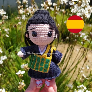 May include: Crocheted amigurumi doll with black hair, a pink face, and large eyes. The doll is wearing a blue and green outfit and is holding a yellow and green bag. The doll is standing in front of a white flower and a red and yellow circle with the Spanish flag.