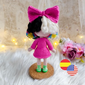 Könnte beinhalten: Handgemachte Häkelpuppe mit einem leuchtend rosa Kleid und einer großen passenden Schleife. Die Puppe hat schwarz-weißes Haar mit bunten Akzenten und grün-weiß gestreifte Schuhe. Die Puppe steht auf einem Korksockel.
