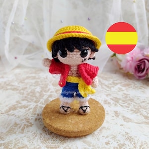 Peut inclure: Figurines amigurumi au crochet d'un personnage de dessin animé portant une chemise rouge, un short bleu, un chapeau de paille jaune et des sandales. Le personnage a les cheveux noirs et de grands yeux. La figurine est posée sur une base en bois brun.
