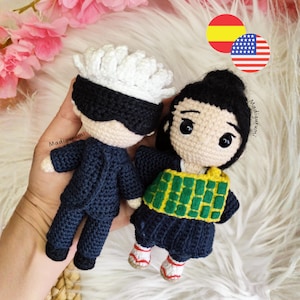 May include: Two handmade crochet dolls. One doll is wearing a dark blue suit with a white hairstyle and a black blindfold. The other doll is wearing a dark blue outfit with a green and yellow design.