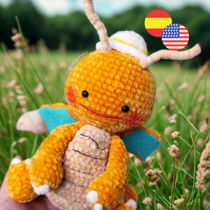 May include: A handmade, plush, orange and tan crochet creature with a white hat and blue wings. The creature has black eyes, a red smile, and white accents on its feet. The background is a field of green grass and wildflowers.