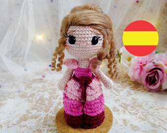 Pode incluir: Uma boneca de crochê com cabelo castanho, vestida com um vestido rosa e bordô, em pé sobre uma base de madeira. A boneca tem grandes olhos pretos e uma expressão doce. Uma bandeira vermelha e amarela é visível no fundo.