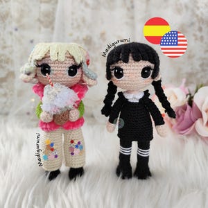 May include: Two handmade crochet dolls with large black eyes. The left doll has blonde hair, a pink jacket, and beige pants with colorful star patches, holding a drink. The right doll has black braided hair and a black dress with white collar.