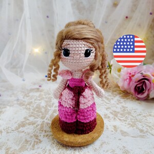 Puede incluir: Una muñeca de crochet con cabello castaño, vestida con un vestido rosa y burdeos, de pie sobre una base de madera. La muñeca tiene grandes ojos negros y una expresión dulce.