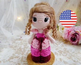 Pode incluir: Uma boneca de crochê com cabelo castanho, vestindo um vestido rosa e bordô, em pé sobre uma base de madeira. A boneca tem grandes olhos pretos e uma expressão doce.