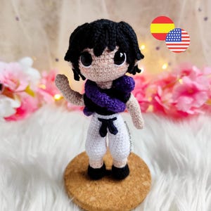 May include: A handmade crochet doll of a character with black hair, large eyes, and a purple scarf. The doll wears a white outfit with a black belt and shoes. It stands on a cork base, with a soft, white background and pink flowers.