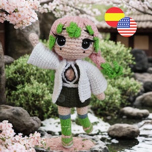 May include: A crocheted doll with pink and green hair, large black eyes, and long eyelashes. The doll wears a white jacket over a dark top, a dark skirt, and green socks with pink accents. One arm is raised, and the doll stands on a base.