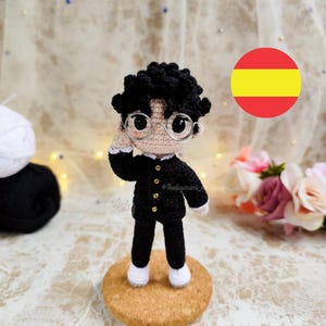 May include: A handmade amigurumi doll of a person wearing a black school uniform with gold buttons, round glasses, and white shoes. The doll has black curly hair and is standing on a cork base. The doll is crocheted.
