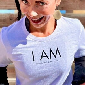 Peut inclure: Une personne portant un t-shirt blanc avec le texte "I AM the little engine that could." La personne est assise sur un tabouret noir devant un mur en bois.