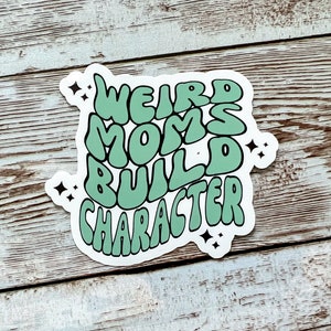Könnte beinhalten: Ein grüner Aufkleber mit dem Text "Weird Moms Build Character" in einem Retro-Schriftart-Stil. Der Aufkleber hat einen weißen Rand und ein paar kleine weiße Sterne um den Text herum.