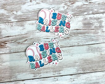 Op de afbeelding: Twee stickers met een honkbal en de tekst "Busy Raising Ballers" in een retro-lettertype. De stickers zijn op een witte houten achtergrond. De tekst is in verschillende kleuren, waaronder roze, blauw en groen.
