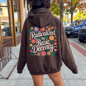 Peut inclure: Sweat à capuche chocolat foncé avec l'inscription "Radicalized by Basic Decency" dans un motif floral au dos. Le sweat à capuche est de couleur unie, avec une capuche et des manches longues. La photo est prise en extérieur.