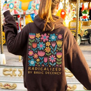 May include: A dark chocolate-coloured hoodie with a floral design on the back. The design features colourful flowers and the text "RADICALIZED BY BASIC DECENCY". The hoodie has a hood and long sleeves.