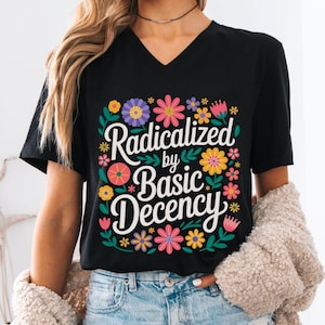 May include: Black V-neck t-shirt with the text "Radicalized by Basic Decency" in white, surrounded by colorful flowers. The shirt is a casual wear item.