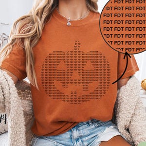 May include: An orange t-shirt with a graphic design made of the letters "FDT" repeated. The shirt is paired with denim shorts and a cream-colored cardigan. A small pumpkin and a woven basket are in the background.