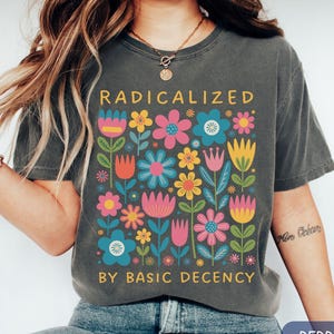 May include: Dark gray t-shirt with the text "RADICALIZED BY BASIC DECENCY" in yellow. The shirt features a colorful floral design with pink, blue, yellow, and orange flowers. The shirt is a casual style.