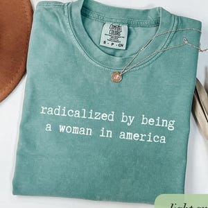 May include: Light green t-shirt with the text "radicalized by being a woman in america" in white. A silver necklace with a pendant rests on the shirt. The shirt is a Comfort Colors brand.