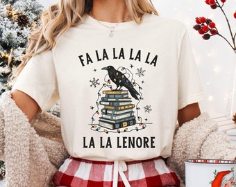 May include: A white t-shirt with a black raven perched on a stack of books. The raven is surrounded by Christmas lights and snowflakes. The text on the shirt reads "Fa La La La La La La Lenore".