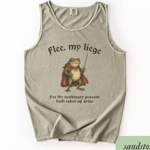 Peut inclure: Débardeur couleur sable avec un dessin humoristique d'une grenouille en cape tenant une épée. Le texte dit "flee, my liege" au-dessus de la grenouille et "for the nonbinary peasant hath taken up arms" en dessous.