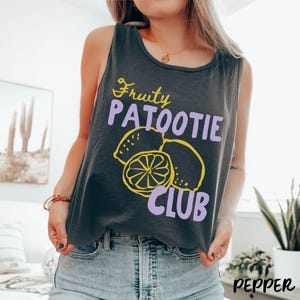 May include: A charcoal grey sleeveless tank top featuring a graphic print with the yellow text Fruity, the purple text PATOOTIE, and the yellow text CLUB, accompanied by a yellow line-art illustration of three lemons.