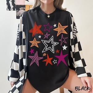 May include: Black long-sleeve shirt with a colorful star graphic. The stars are in shades of orange, pink, purple, and white. A black and white checkered cardigan is worn over the shirt. The word "BLACK" is printed at the bottom.