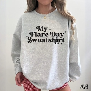 May include: A light gray crewneck sweatshirt featuring the black text My Flare Day Sweatshirt centered on the chest. The text is surrounded by small four-pointed star icons. The garment has long sleeves and a ribbed collar, cuffs, and hem.
