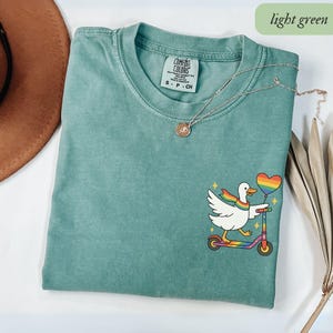 May include: Light green Comfort Colors t-shirt with a graphic of a white duck riding a rainbow scooter and holding a rainbow heart balloon. The duck wears a rainbow scarf. A silver necklace with a pendant is also visible.