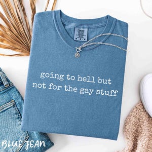 May include: A blue t-shirt with the text "going to hell but not for the gay stuff" in white. A silver necklace with a circular pendant is draped across the shirt. The shirt is folded and placed on a white surface.