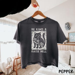 May include: Dark gray t-shirt with a white graphic of a cat surrounded by flowers, and the text "BE KIND & RAISE HELL". The shirt is hanging on a wooden hanger.