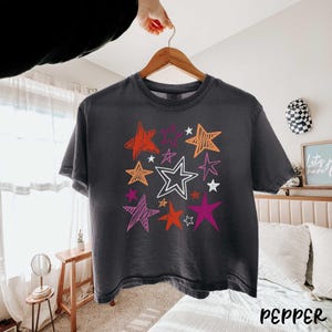May include: Dark gray t-shirt with a colorful star design. The stars are in shades of orange, red, pink, and white. The word "pepper" is printed in white at the bottom right corner of the shirt.