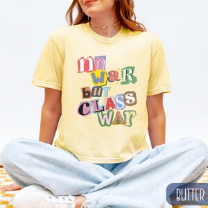 May include: A light yellow t-shirt with the text "no war but class war" in a colorful, cut-out style. The shirt is short-sleeved and has a relaxed fit. The model is wearing light blue jeans.