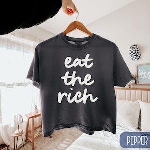 May include: A dark gray t-shirt with the words "eat the rich" in white script. The shirt is hanging on a wooden hanger. The word "pepper" is in a blue rectangle in the bottom right corner.