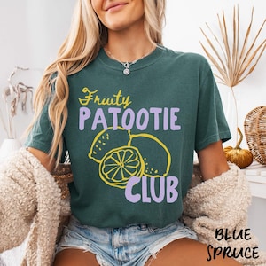 May include: A teal-colored t-shirt with the words "Fruity Patootie Club" in yellow and purple. The shirt features a lemon graphic. The words "Blue Spruce" are printed in the bottom right corner.