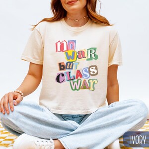 May include: Ivory-colored t-shirt with the text "no war but class war" in a colorful, cut-out style. The shirt is short-sleeved and has a relaxed fit. The model is wearing light blue jeans.