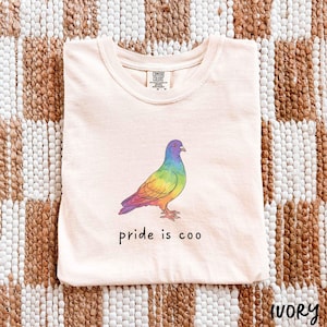 May include: Ivory-coloured t-shirt with a colourful illustration of a pigeon with rainbow-coloured feathers. The text "pride is coo" is printed below the bird. The shirt is made of a soft material.