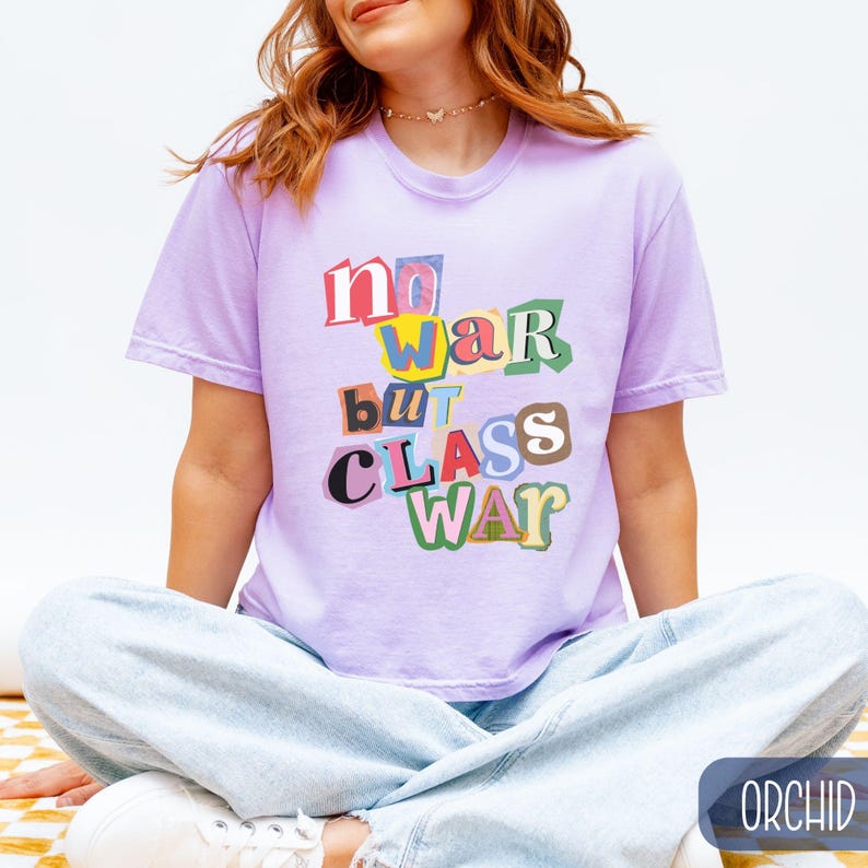 May include: A light purple t-shirt with the text "No War But Class War" in a colorful, cut-out style. The shirt is short-sleeved and has a relaxed fit. The model is wearing light blue jeans.