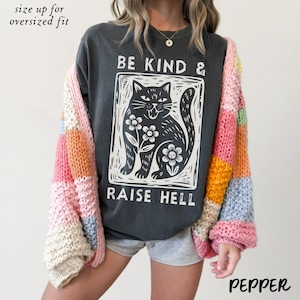 May include: Dark gray sweatshirt with a black and white cat graphic that reads "BE KIND & RAISE HELL." A colorful patchwork cardigan in pink, orange, yellow, and blue is worn over the sweatshirt. The text "size up for oversized fit" is at the top.